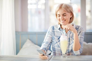 Kafeteryada dinlenirken lezzetli kokteyl ve akıllı telefon mesajlaşması ile gülümseyen genç kadın