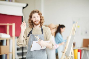 Not defteri ve fırçaları olan genç ressam sanat stüdyosunda duruyor ve soru sormak için elini kaldırıyor.