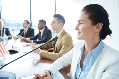 Young elegant female delegate sitting by table with microphone in front and speaking at conference clipart