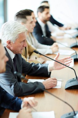 Mature grey-haired delegate speaking in microphone while commenting speech of one of participants of summit clipart