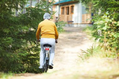 Sarı kazak kır evine kadar bisiklet sürme gri saçlı yaşlı adamın arka görünümü, kaba yolda aktif kıdemli adam bisiklet