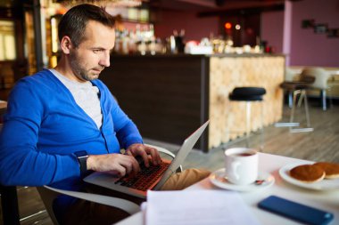 İçerik girişimci yakışıklı erkek serbest mavi hırka masada oturan ve kahvaltı sırasında kafede iş yaparken dizlerinin üzerinde dizüstü tutmak
