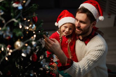 Genç adam ve küçük kızı Noel ağacını oyuncaklar ve çelenklerle süslüyorlar.