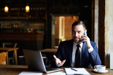 Resmi ceket li içerikten emin yakışıklı işadamı restoran masasında otururken ve cep telefonuyla konuşurken ve gesturing yaparken iş sorunlarını çözer