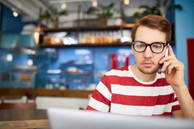 Akıllı telefonu olan genç bir adam bilgisayardaki verileri okurken müşterisine ya da iş arkadaşına telefon ediyor.