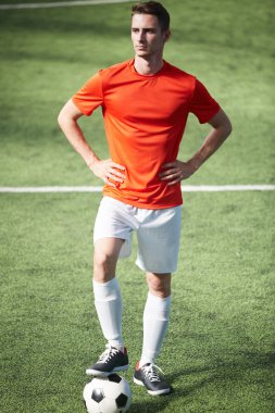 Genç emin futbolcu akimbo içinde poz topu bir ayağı yeşil alan üzerinde duran