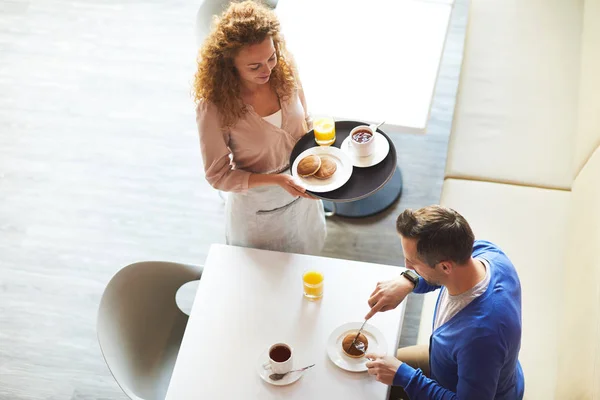 新鮮なパンケーキとトレイを保持若いウェイトレス ビジネスマンは彼の朝食を食べるテーブルのそばに立っている間茶とオレンジジュースのガラスのカップ — ストック写真