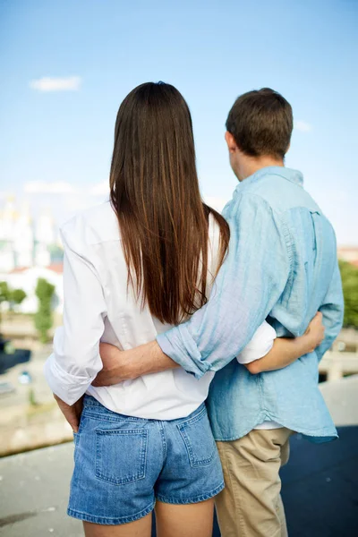 年轻夫妇穿着休闲服装互相拥抱 在屋顶上欣赏城市景观的同时 互相看距离的后景 — 图库照片