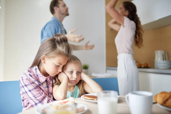 両親が背景に議論している間 台所のテーブルのそばに座っている2人の悲しい女の子 — ストック写真