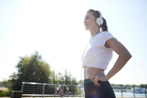 有線ヘッドフォンの新しい成果について考えている間距離にして屋外で立って笑顔の楽観的な若い女性 — ストック写真