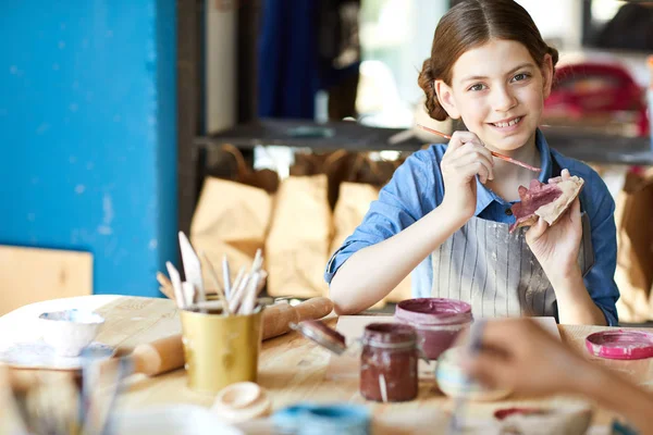 美術工芸のレッスンに座りながら 自作粘土のおもちゃを描く絵筆を持つ幸せな女の子 — ストック写真