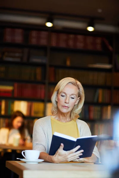 金发碧眼的女性阅读书在蓝色封面 而花费时间在咖啡馆或图书馆 — 图库照片
