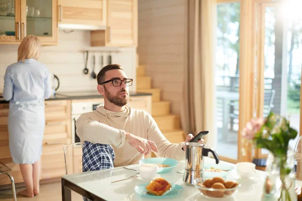 年轻的留胡子的白种人坐在厨房的桌旁 看着电视震惊的表情 而他的妻子正在做早餐 — 图库照片