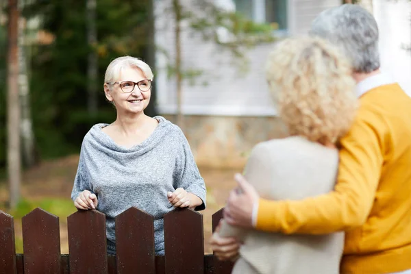 金发成熟的女人在眼镜和灰色的拉过看着她的邻居在交谈通过围栏 — 图库照片