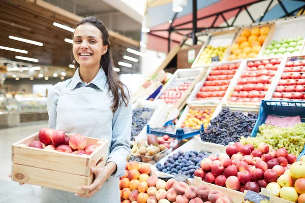 欢快的年轻女杂货零售商在围裙站在食品摊位和拿着一盒苹果 她卖新鲜水果和蔬菜 — 图库照片