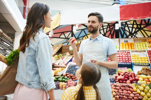 Content handsome young grocer in apron talking to customer and recommending to buy peach while selling fresh food at farmers market