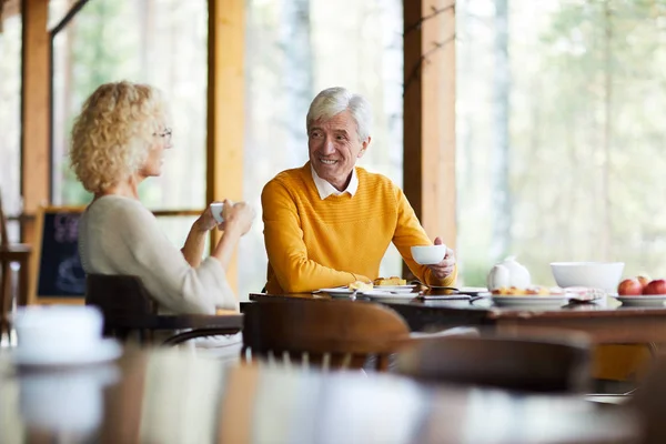 黄色のプルオーバーの成熟した男と彼のブロンドの妻はテーブルのそばに座って デザートとお茶を飲んで カフェで話しています — ストック写真