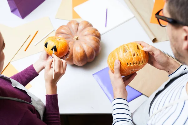 Close-up of busy colleagues standing at table and making design for Halloween pumpkins, man carving pumpkin while woman painting small pumpkin in studio