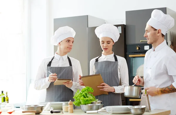 Pensive young students in chefs hats and aprons asking questions to chef with cooking pan while studying at master class