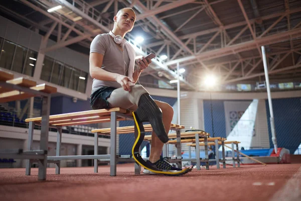 年轻的沉思运动员 右腿截肢 在智能手机上滚动 而在训练后在长凳上休息 — 图库照片
