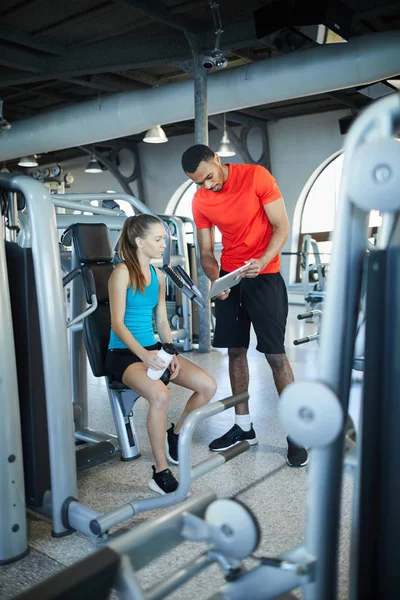 Active female watching online sports video in touchpad held by her trainer in fitness center at break