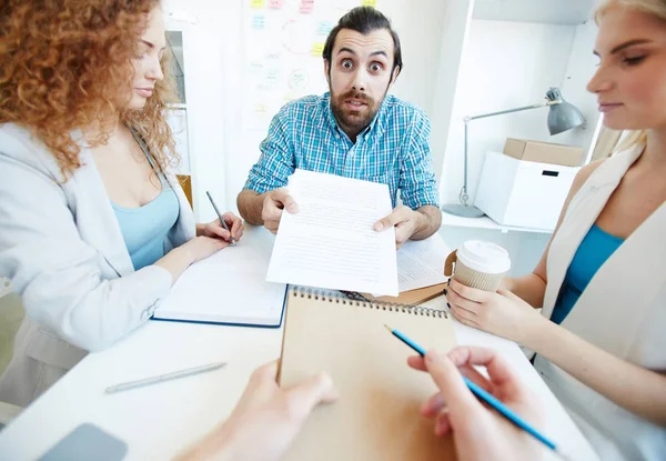 Annoyed businessman with document looking at colleague or subordinate sitting in front during their conversation