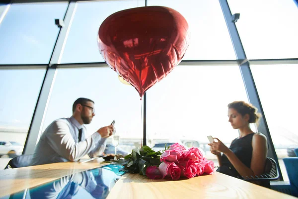 バラと赤いハート形の風船の束と彼らのガジェットを使用して サーブテーブルに座っているバレンタインのカップル — ストック写真