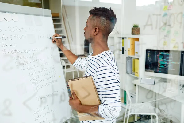 Young trainee with notepad writing down software codes on whiteboard in office