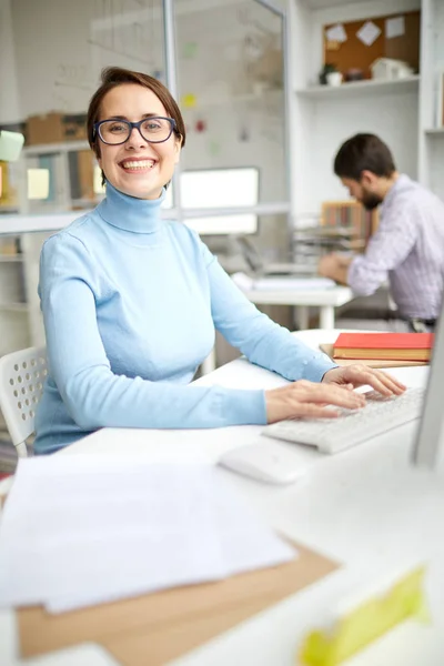 オフィスの机でコンピュータモニターの前で働きながら 歯ごたえのある笑顔で元気なマネージャー — ストック写真