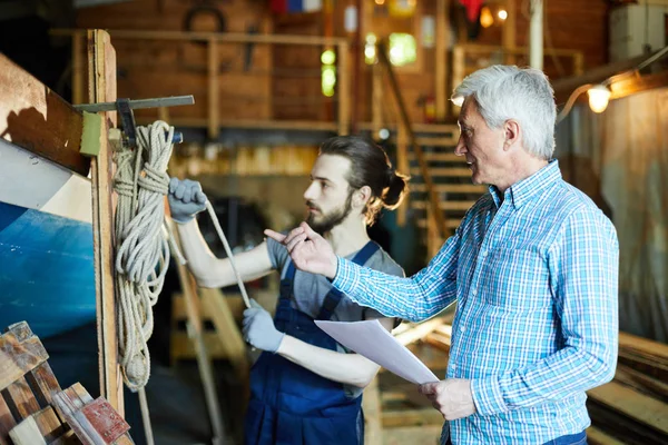 Aged engineer or master pointing at vessel and talking to his young colleague in workshop