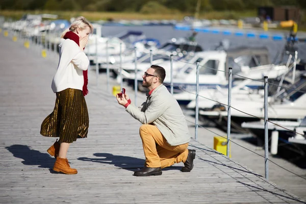 スタイリッシュなひげを生やした白人の男性は 彼の膝の上に立って 桟橋で彼のガールフレンドに提案し 婚約指輪でボックスを保持し 幸せに微笑んで — ストック写真
