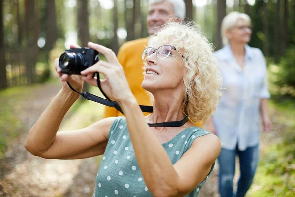 微笑兴奋的成熟女士与卷发戴眼镜抬头 拍摄动物在森林中 而与朋友散步 — 图库照片