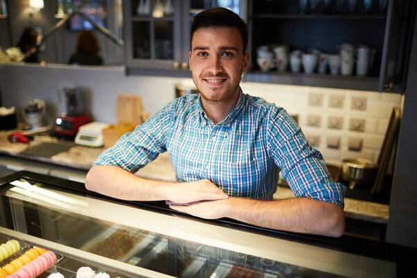 Young seller looking at camera while leaning by large display with desserts and pastry in cafe