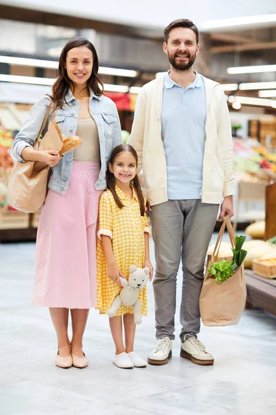 スーパーマーケットに立って カメラを見てカジュアルな衣装で美しい若い家族を笑顔 彼らは一緒に買い物をしています — ストック写真