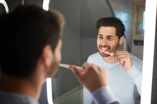 陽気なハンサムな若いひげの男は バスルームで照らされた鏡を見て 朝に歯ブラシで歯をきれいに — ストック写真