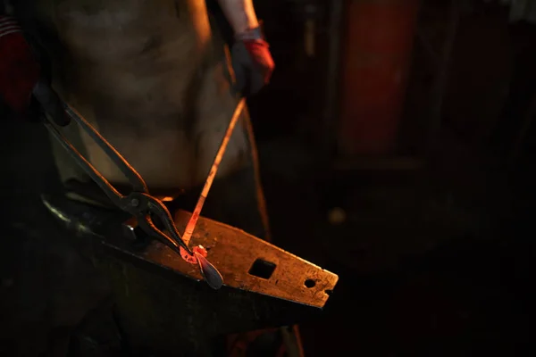 暗いワークショップでアンビルで働いている間 トングで加熱された金属板を形作るエプロンで認識できない鍛冶屋のクローズアップ — ストック写真