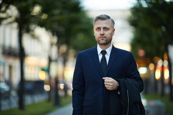 都会の環境の中を歩きながらコートを持つエレガントなスーツを着た成熟した真面目なビジネスマン — ストック写真