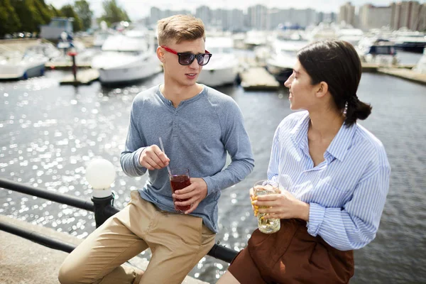 一对年轻夫妇 喝着饮料 坐在篱笆上 身后是水边 谈着话 — 图库照片