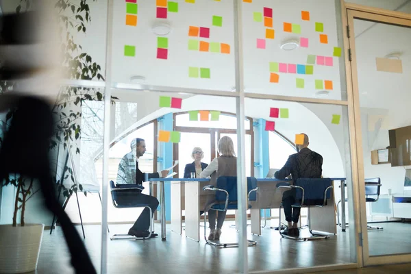 Team of contemporary employees sitting by table inside office with transparent walls and organizing work