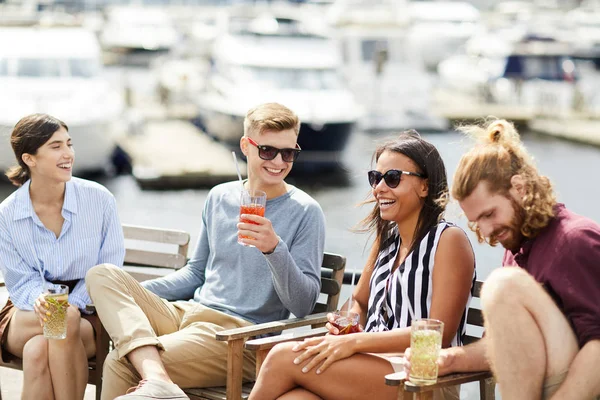 Happy young laughing people with drinks enjoying summer weekend by waterside