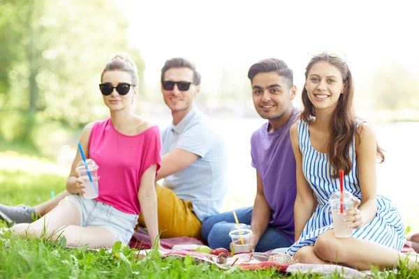 緑の芝生の上に座って カメラを見て 夏休みを楽しんで飲み物を持つ2人の幸せな若いカップル — ストック写真