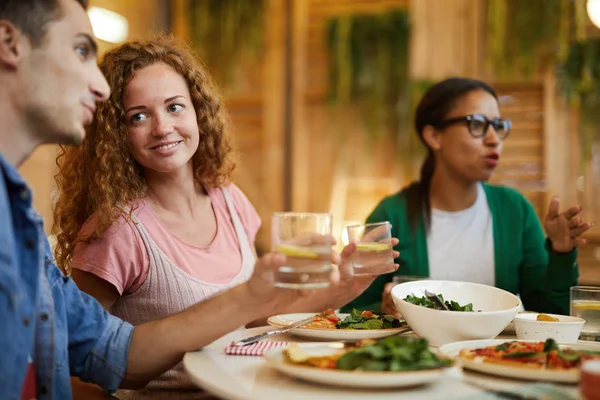 微笑的女孩与波浪头发看着她的男朋友在与朋友的晚餐服务表交谈 — 图库照片