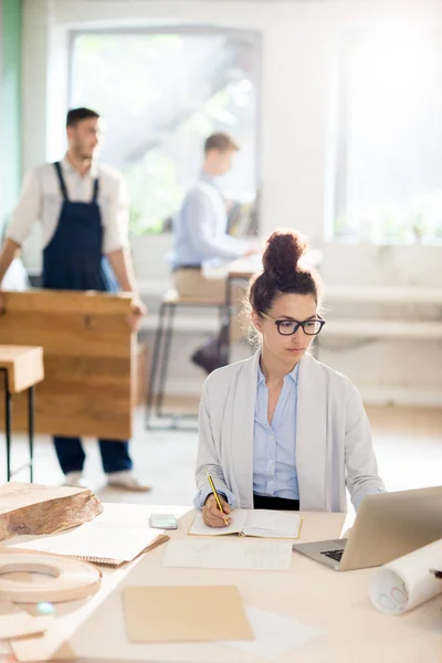 Young female analyst sitting in front of laptop and solving organization moments in working environment