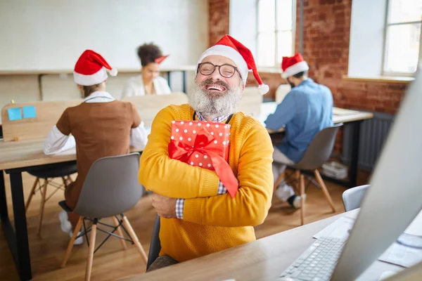 オフィスの職場で座っている間にプレゼントとプレゼントボックスを抱きしめるサンタキャップの興奮したシニアビジネスマン — ストック写真