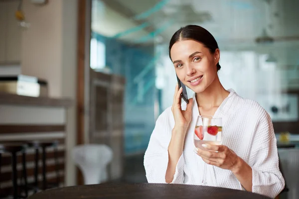 Young smiling woman talking by smartphone while spending time in cafe and having light cocktail