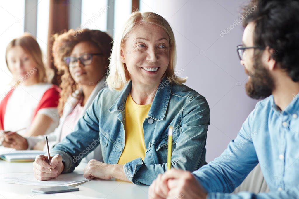 Happy mature businesswoman looking at one of colleagues with smile while listening to his explanation of working point