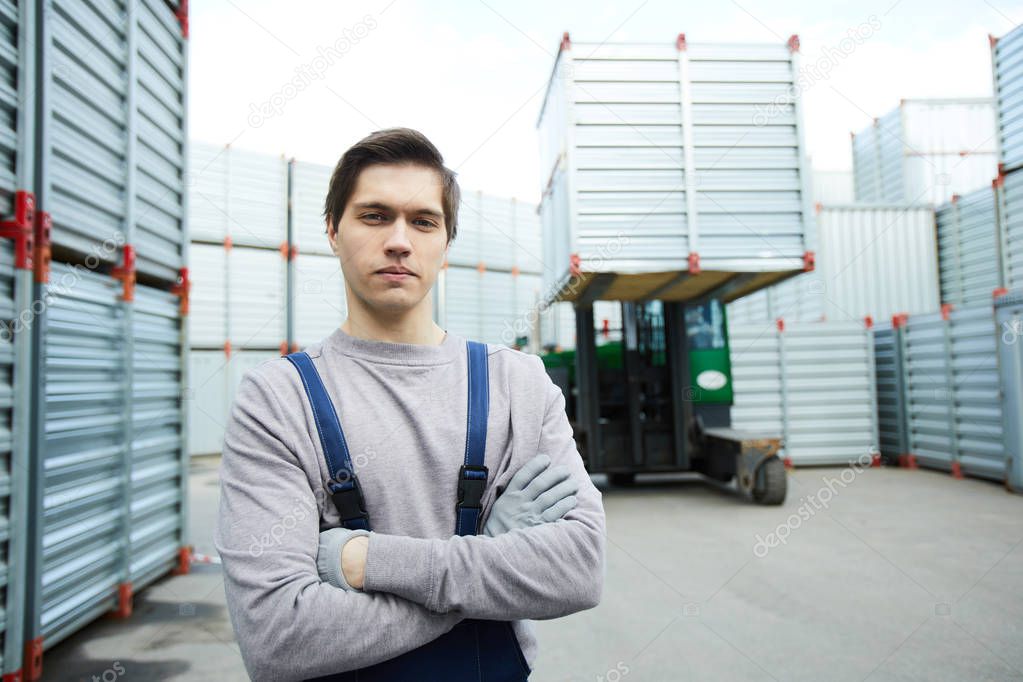 Cross-armed worker in grey pullover and dark blue overalls looking at camera on territory of transportation company