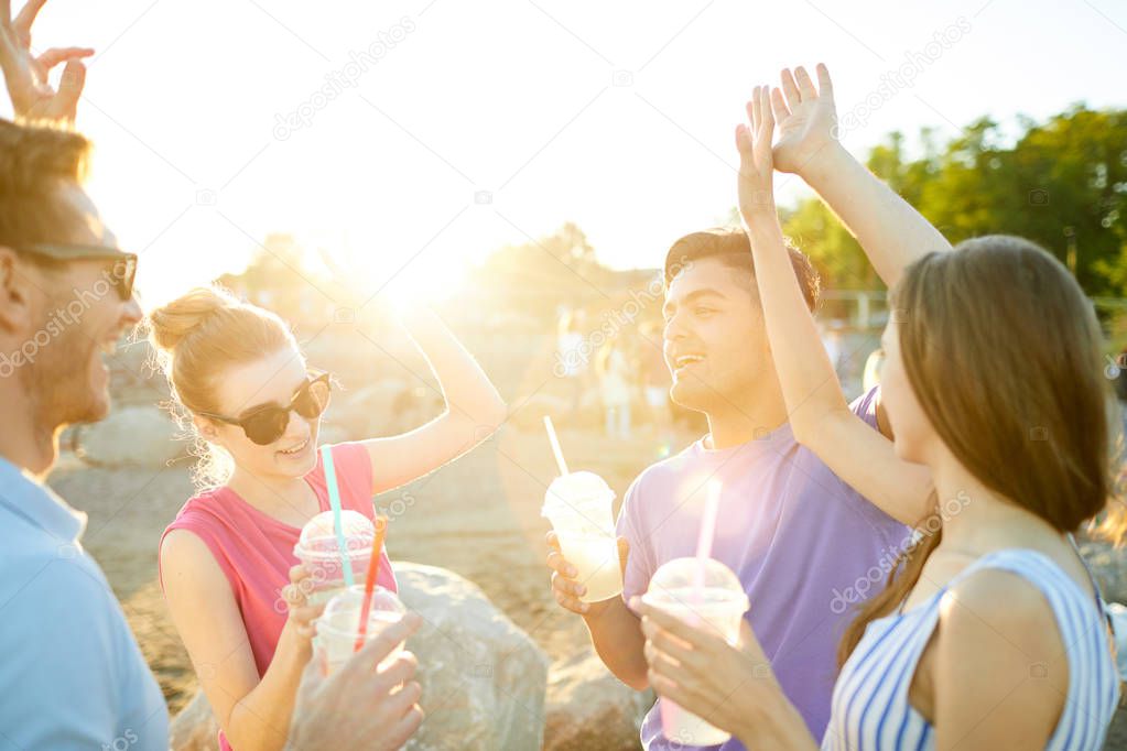 Two excited teenage couples with drinks dancing and singing on the beach on hot summer day