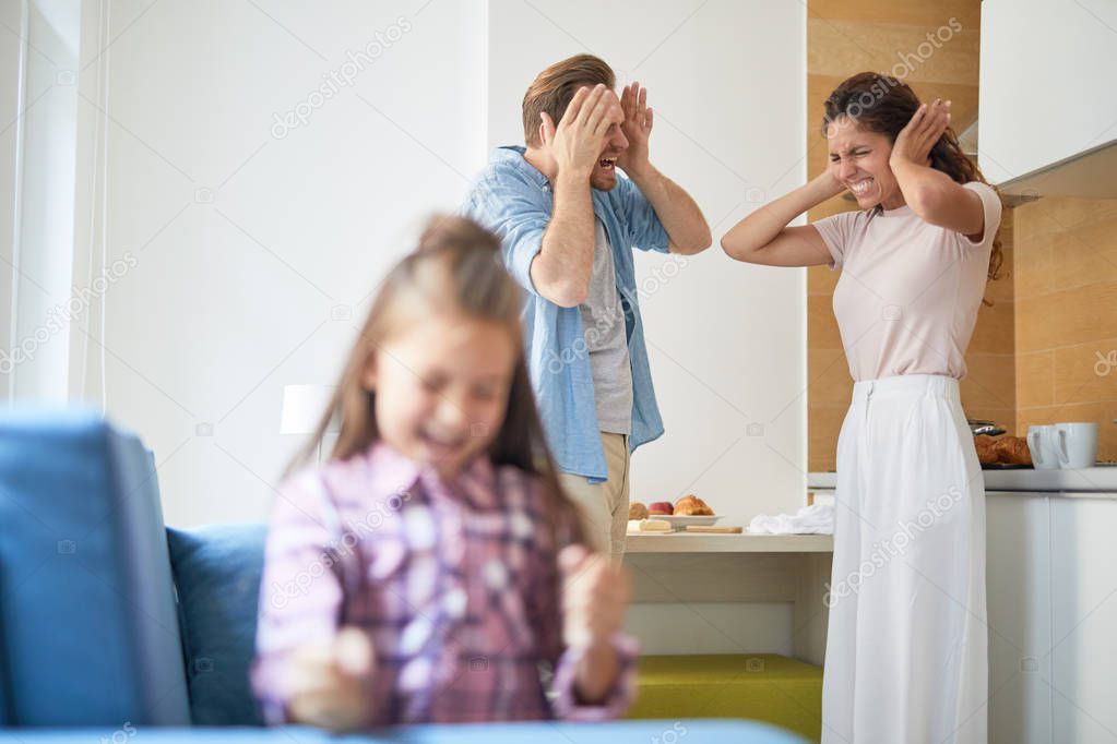 Stressed parents covering their ears in horror while their daughter shouting loudly and playing in armchair