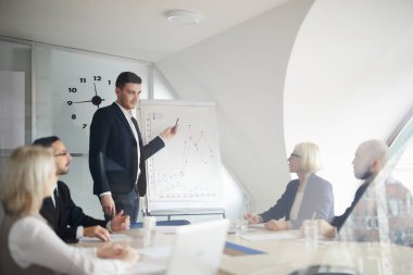 Zarif takım elbiseli genç ekonomist, seminerdeki meslektaşlarına açıklarken tahtadaki grafiklere işaret ediyor.